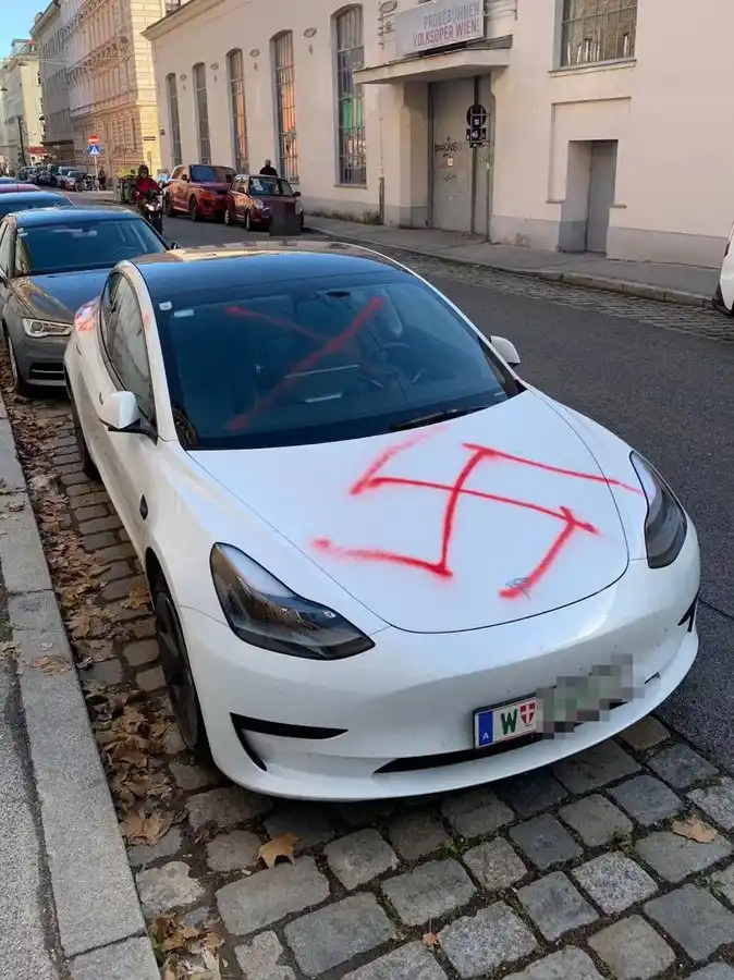 a white tesla model 3 is parked on a cobblestone street with a red swastika spray-painted on its hood and windshield. The…