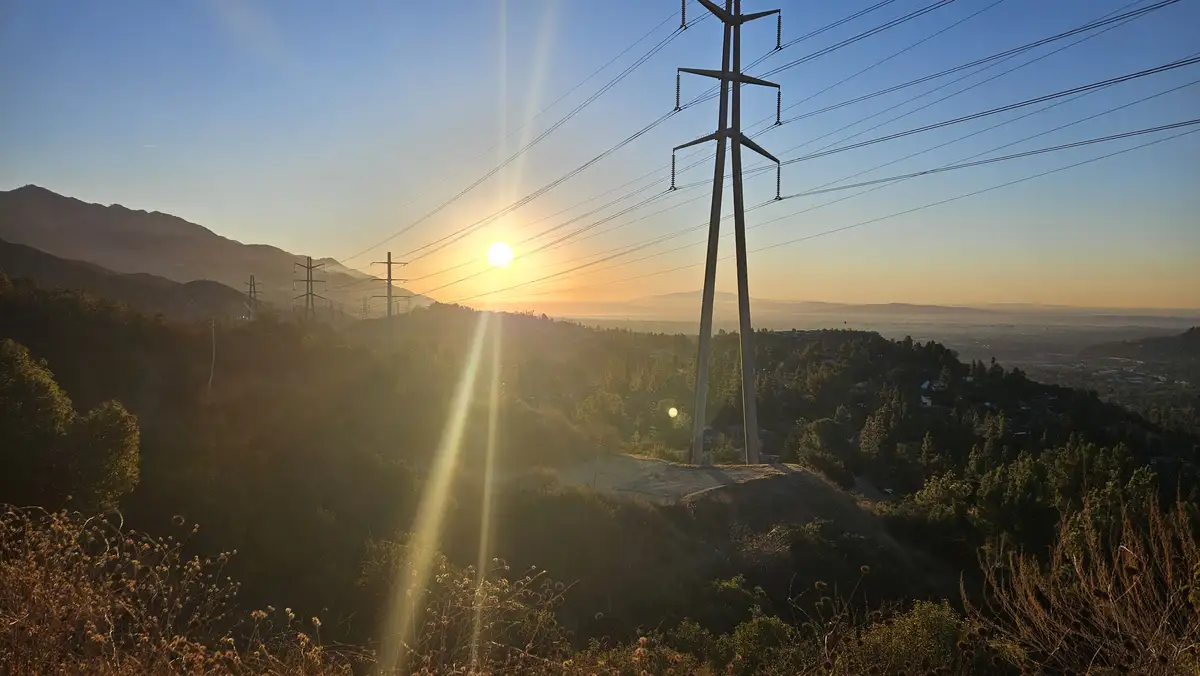 A scenic photo of a sunrise or sunset over rolling hills with power lines and transmission towers silhouetted against the…