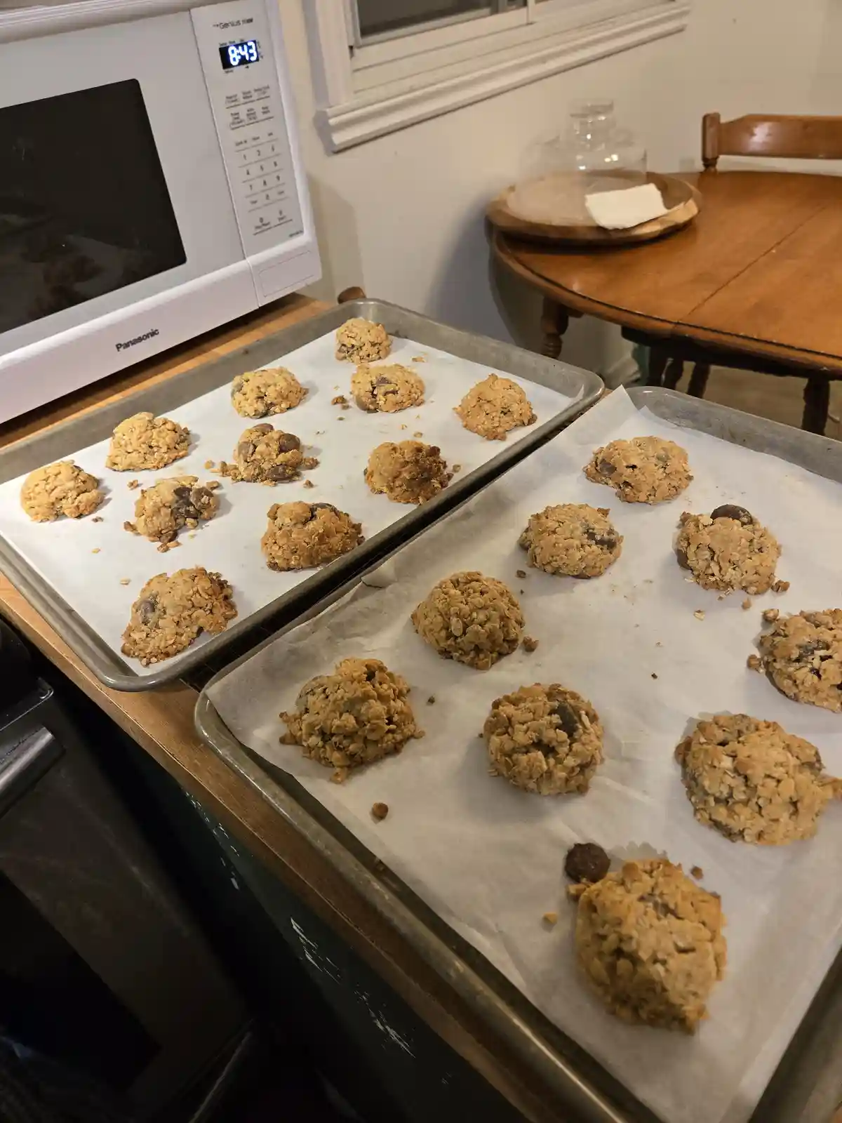 Two baking sheets lined with parchment paper hold freshly baked oatmeal chocolate chip cookies, sitting on a wooden kitchen…