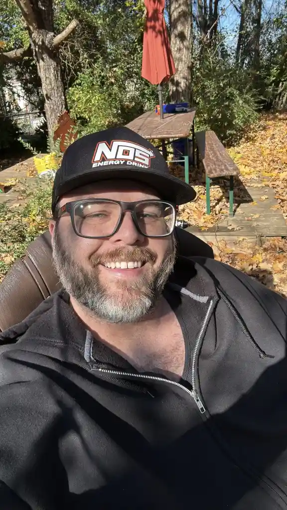 A man with a beard and glasses smiles while wearing a black baseball cap with 'NOS ENERGY DRINK' printed on it. He is…