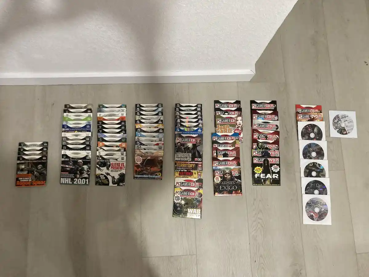 A collection of PC Gamer CD and DVD game discs from the early 2000s are laid out on a light wood floor, arranged in vertical…