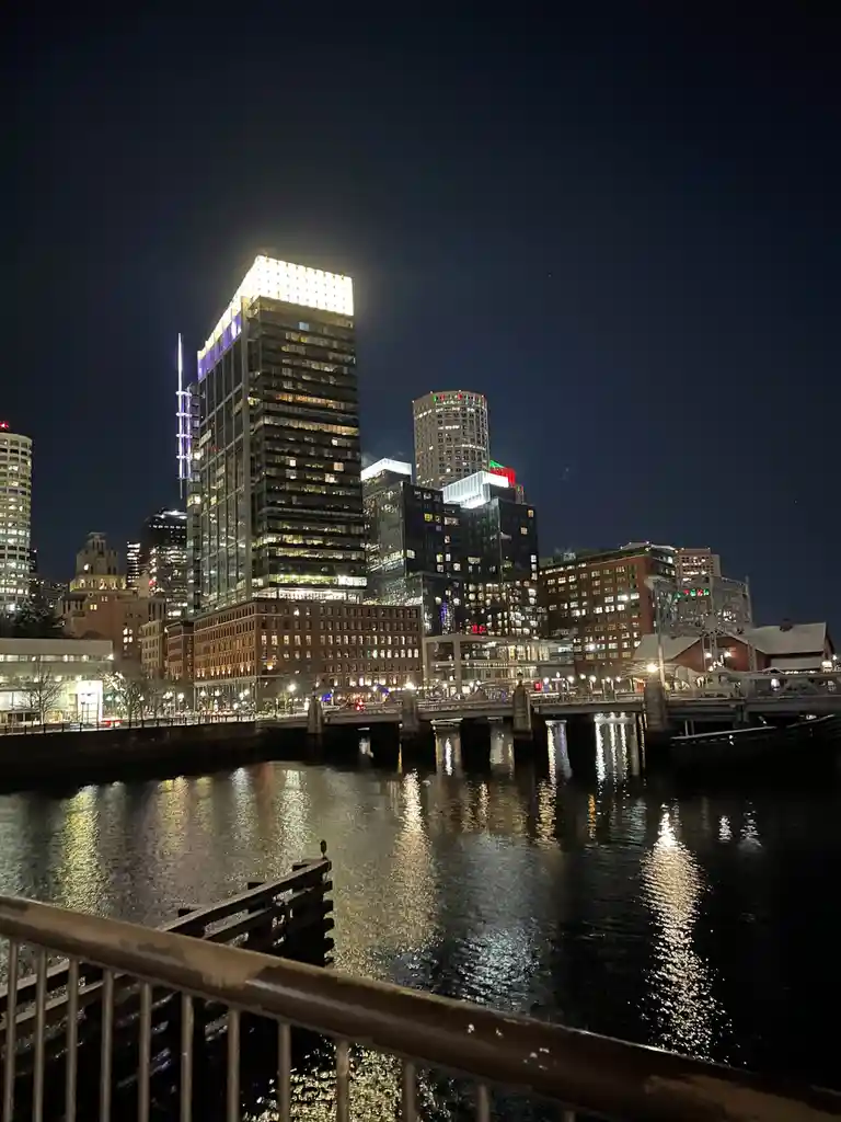 A nighttime cityscape photo showing illuminated skyscrapers and a bridge over a river, with lights reflecting on the water…