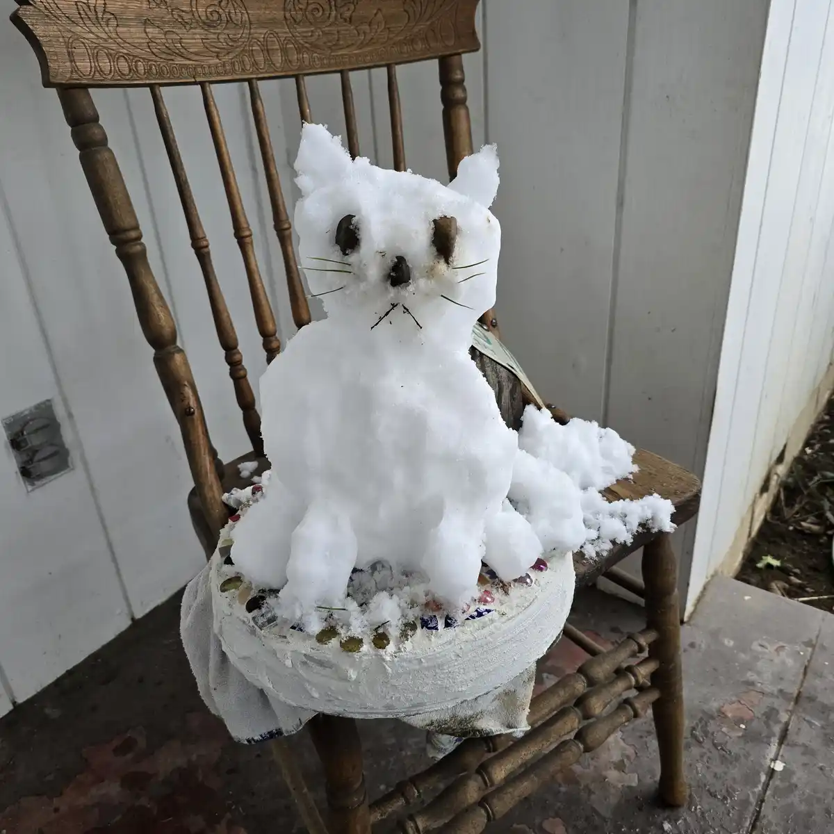 A snow sculpture shaped like a cat sits on a wooden chair outdoors, with small twigs for whiskers and dark objects for eyes…