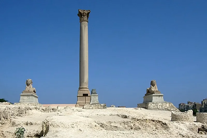 A tall, solitary ancient column stands in the center of a sandy archaeological site under a clear blue sky, flanked by two…