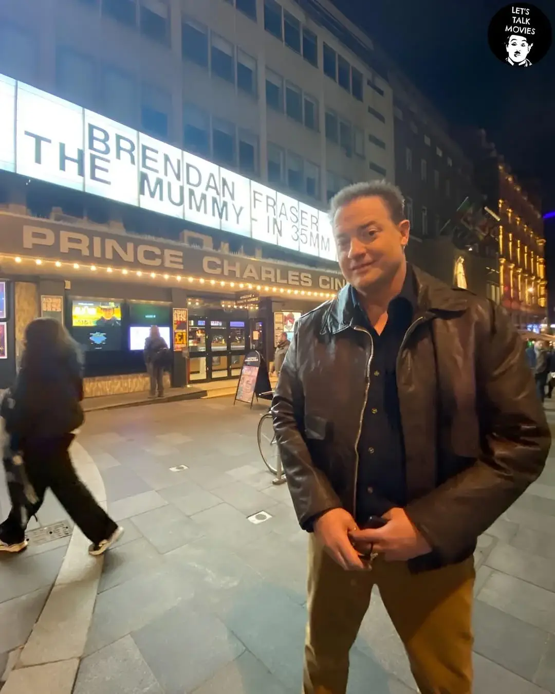 A man in a brown leather jacket stands on a city sidewalk at night in front of the Prince Charles Cinema, which has a lit…