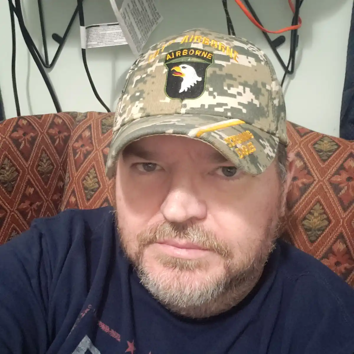 A man with a grizzled beard and mustache stares directly at the camera with a stern, unamused expression while wearing a…