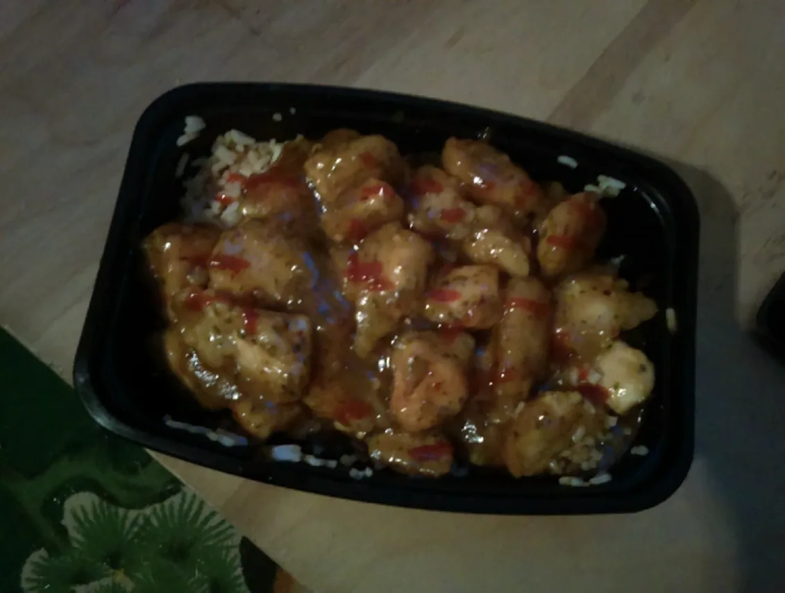 A black takeout container holds chicken pieces covered in a glossy sauce with red specks, sitting atop white rice on a…