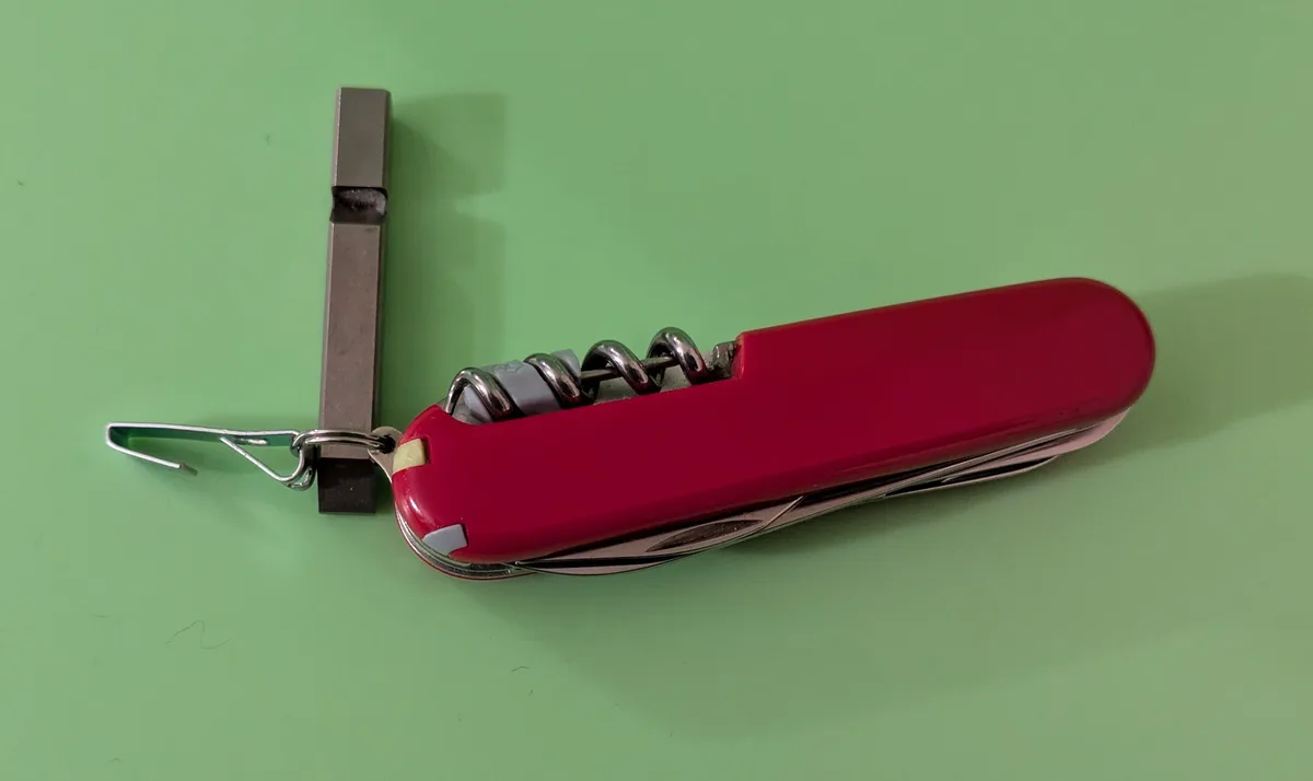 A red Swiss Army knife with its corkscrew tool extended is lying on a green surface, next to a metal tool that looks like a…