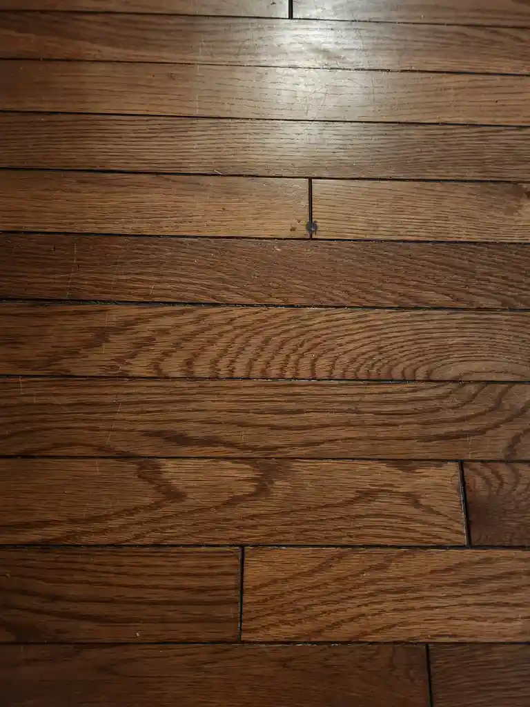 A close-up photo of a worn, dark brown hardwood floor with visible scratches and a small dark stain at a plank seam…