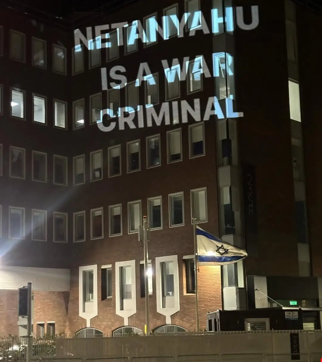 A nighttime photo of a brick building with large white text projected onto its windows reading 'NETANYAHU IS A WAR…