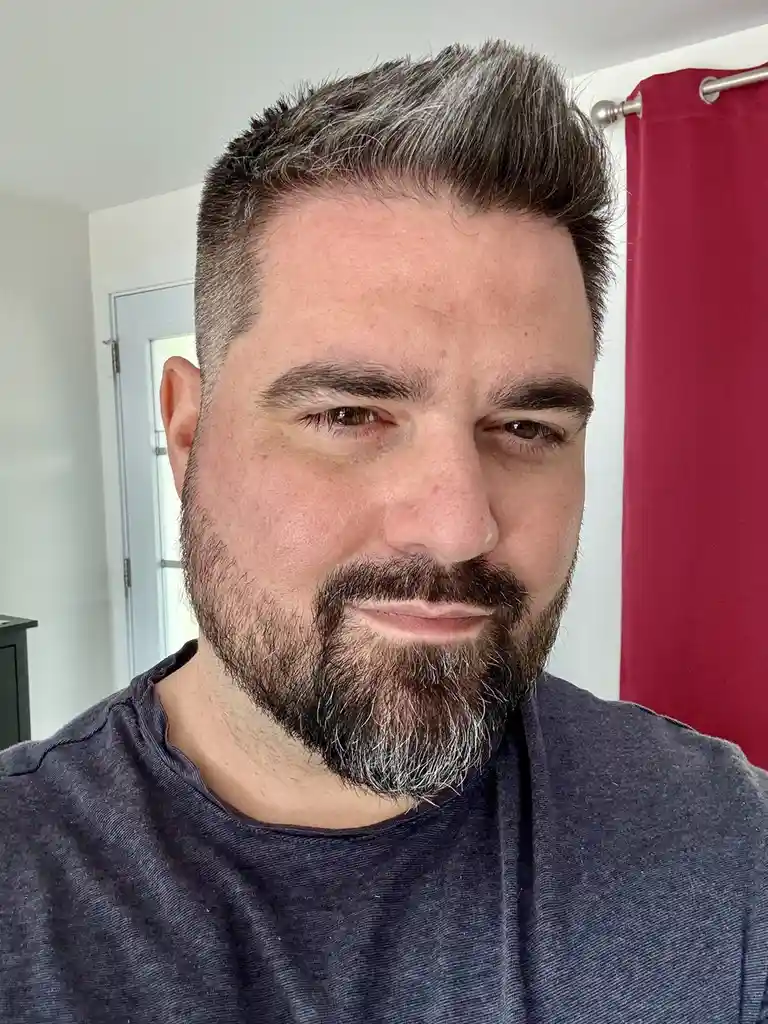 A close-up selfie of a man with a beard and short hair showing visible gray strands, wearing a dark gray t-shirt, looking…