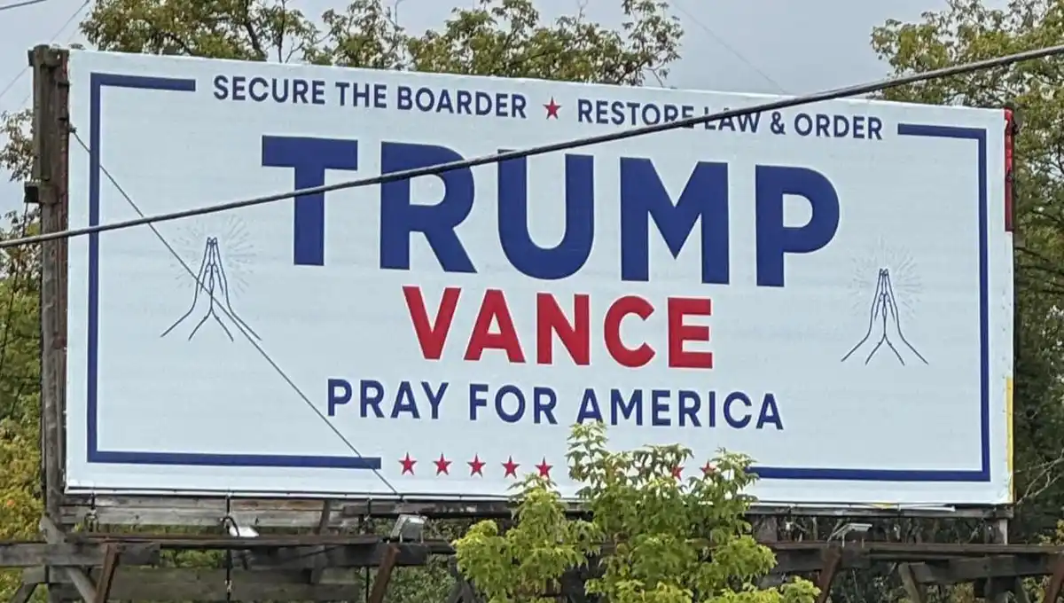 A large outdoor billboard features the names 'TRUMP' in blue and 'VANCE' in red, flanked by illustrations of praying hands…