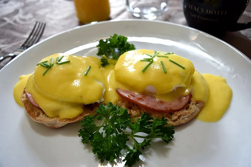 A plate of Eggs Benedict with hollandaise sauce, ham, and chives, garnished with parsley, sits on a table with a fork…