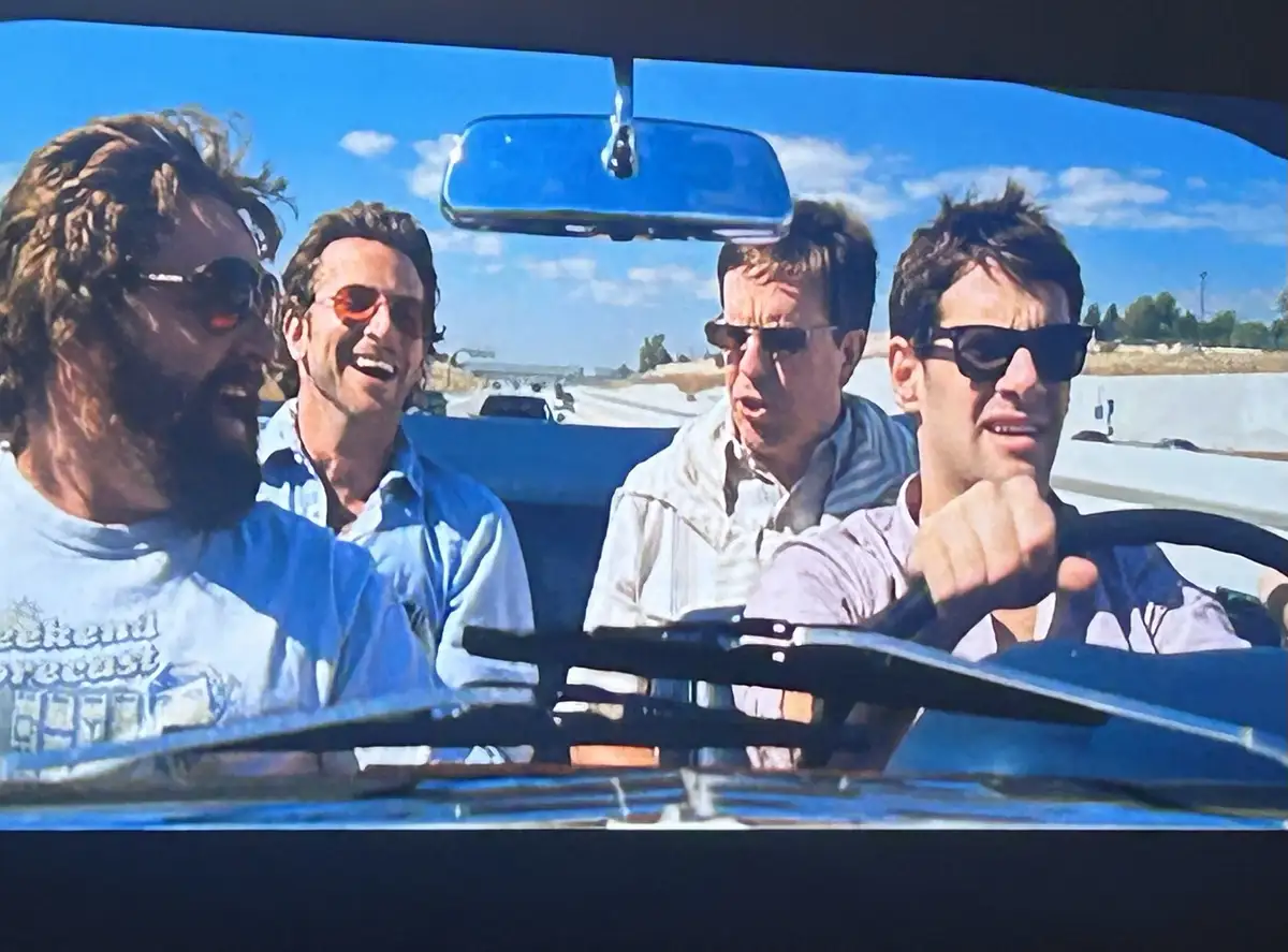 Four men are in a convertible car on a sunny day, with the driver looking stressed and the others laughing. The text overlay…