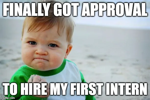 A baby with a smug, satisfied expression stands on a sandy beach, wearing a green and white shirt, holding a small rock. The…
