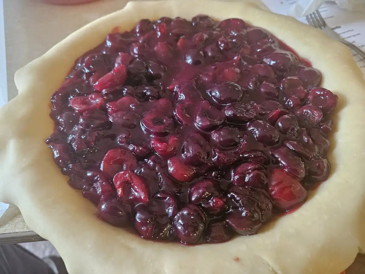 A close-up photo shows a raw pie crust filled with a glossy, dark red cherry filling, with some cherries cut in half. The…