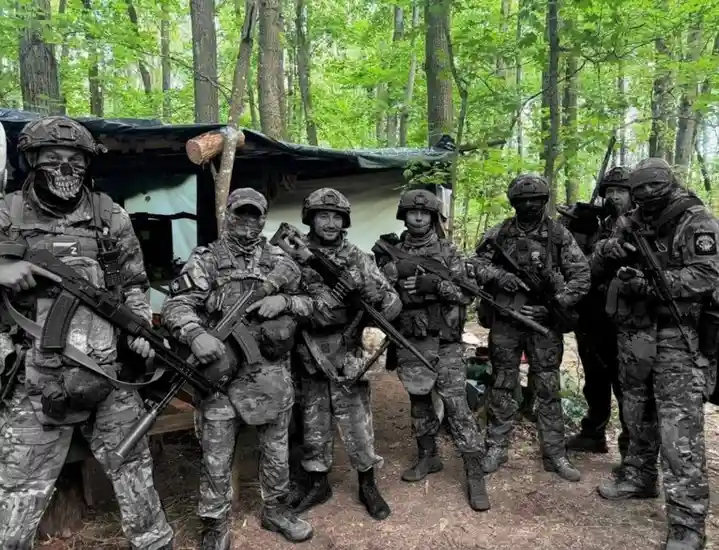 A group of men in full tactical gear, including helmets, camouflage uniforms, and rifles, pose for a photo in a forest…