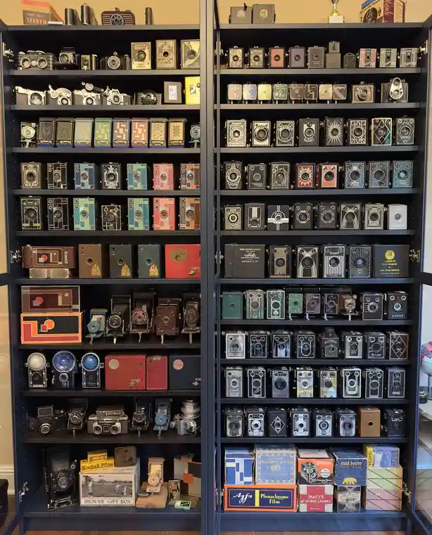 Two open black cabinets are filled with dozens of vintage cameras, neatly arranged on shelves. The cameras vary in size…