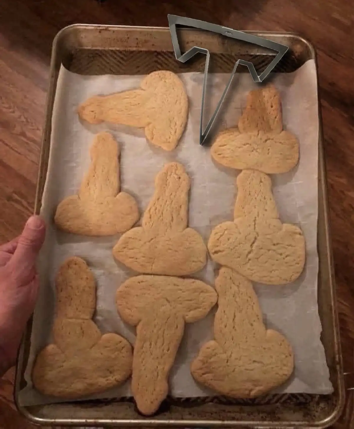 A baking sheet lined with parchment paper holds several golden-brown cookies cut into the shape of a penis. A hand holds the…