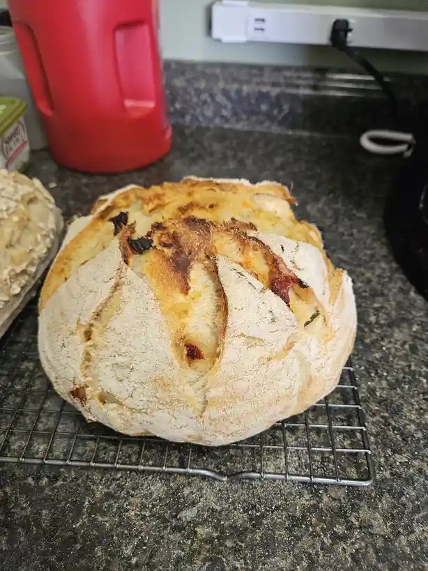 A round, rustic loaf of bread with a golden-brown crust and visible dried tomato pieces sits on a cooling rack on a kitchen…