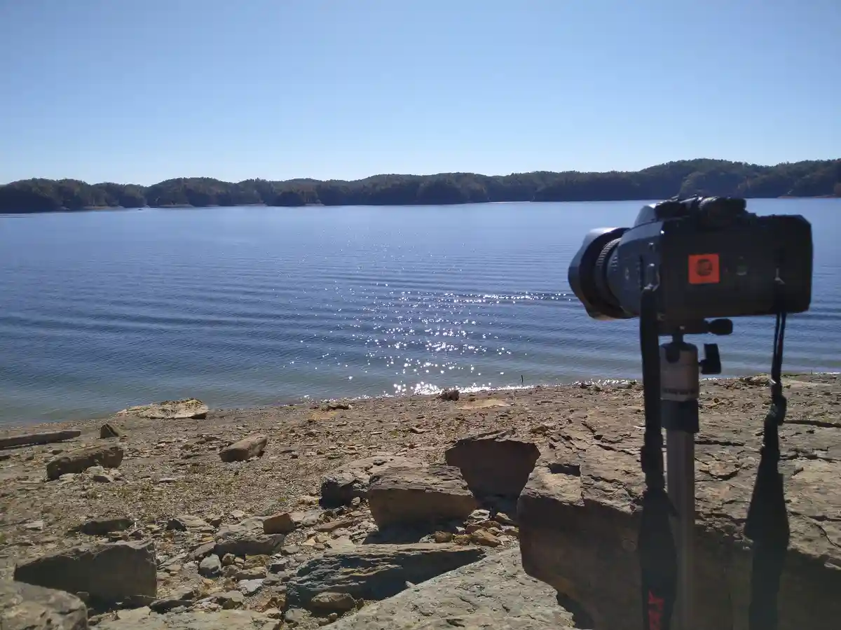 A DSLR camera mounted on a tripod is positioned on a rocky lakeshore, facing a wide expanse of calm blue water under a clear…