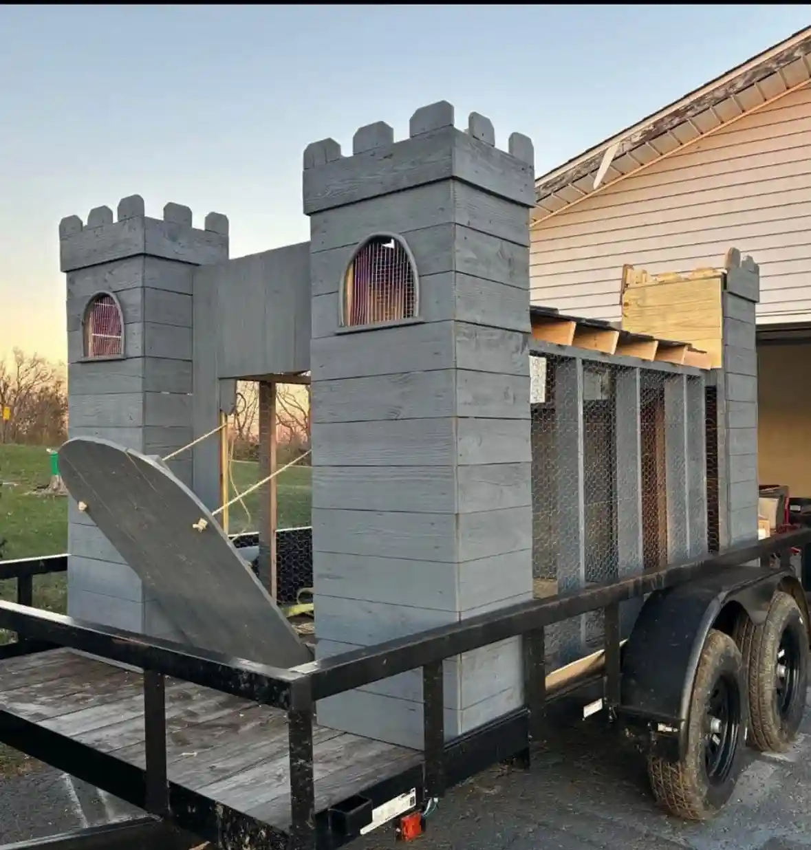 A partially constructed wooden castle with gray-painted planks and crenellated towers sits on a black utility trailer…