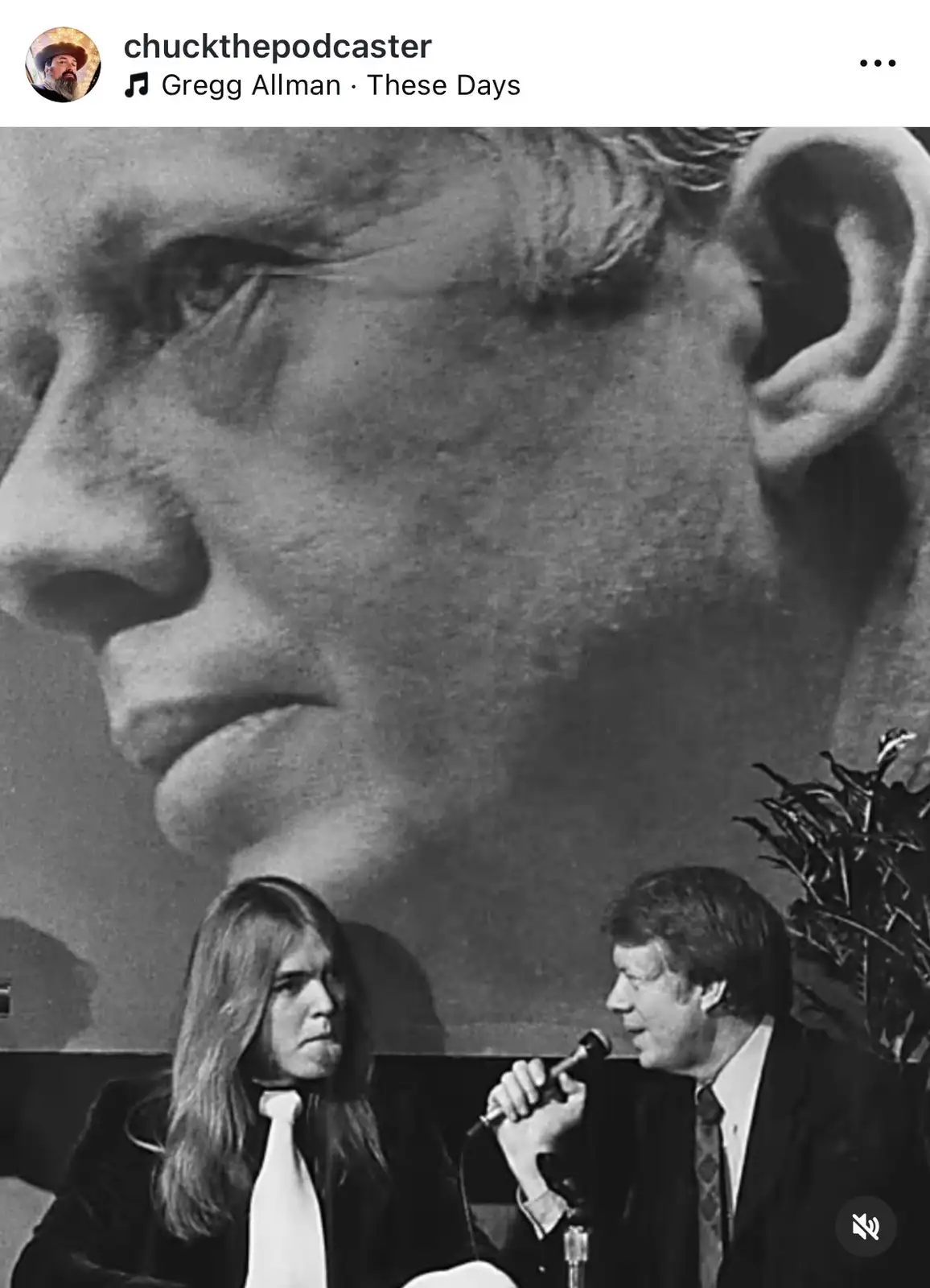 A black-and-white photo shows a man with long hair and a white tie looking unimpressed while sitting next to a man in a suit…