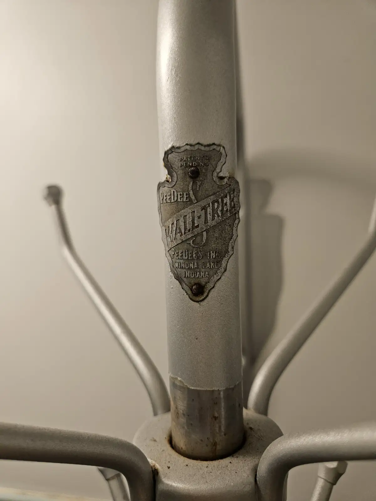 A close-up photo of a silver metal coat rack with a worn, shield-shaped manufacturer's label that reads 'PeeDee Wall-Tree'…
