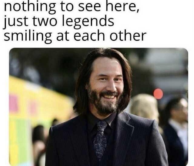 The image shows a man with long dark hair and a beard, wearing a dark suit and tie, smiling broadly at the camera. The…