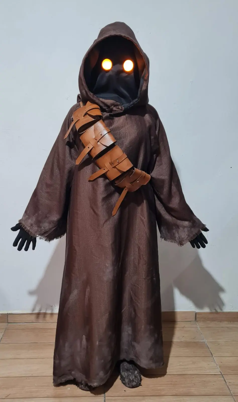 A person in a brown hooded robe with glowing orange eyes and a leather strap across the chest stands against a plain wall…