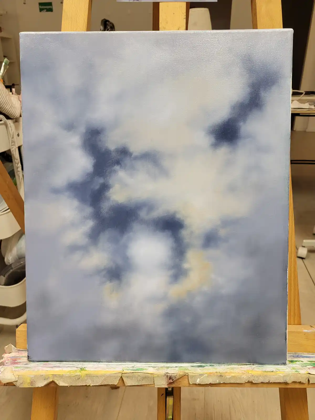 A canvas on an easel displays a painting of a cloudy sky with soft blue and white tones, showing no distinct horizon or sun…