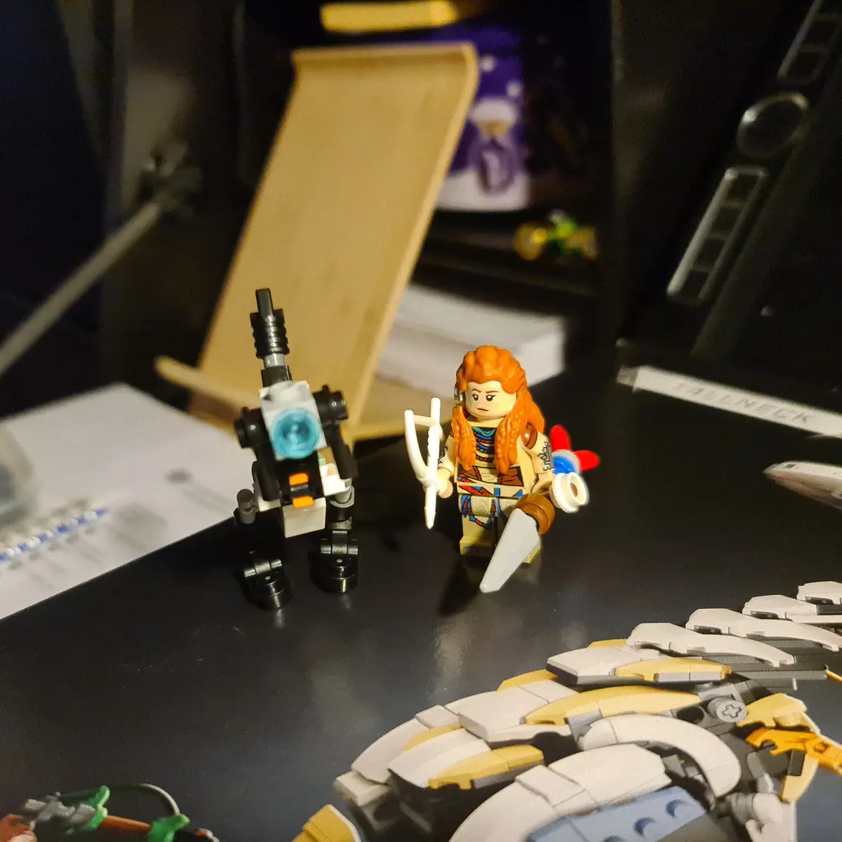 A photo shows two LEGO minifigures on a desk: one is a female warrior with orange hair holding a bow and dagger, and the…