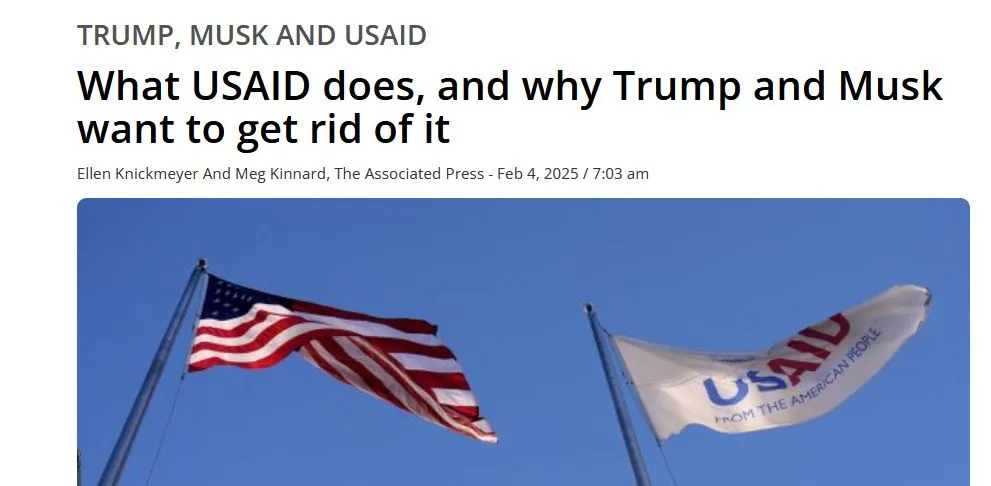 An image shows two flags waving against a clear blue sky: the American flag on the left and a white USAID flag on the right…