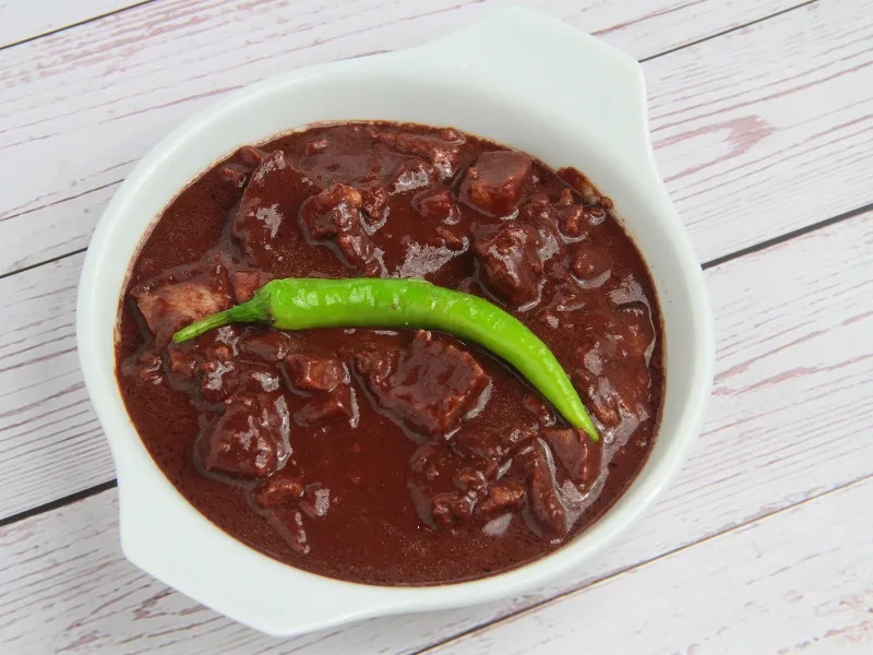 a white bowl filled with a dark brown, thick stew containing chunks of meat, placed on a white wooden surface. A single…