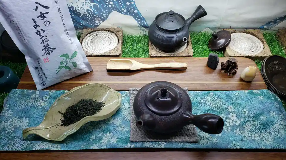 A still life of traditional Japanese tea ceremony items arranged on a wooden table, including two dark ceramic teapots, a…