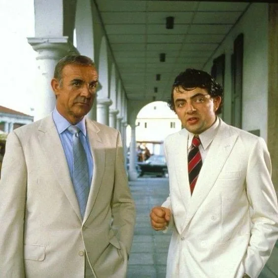 two men in white suits stand side by side under a colonnade; the man on the right has a shocked, wide-eyed expression while…
