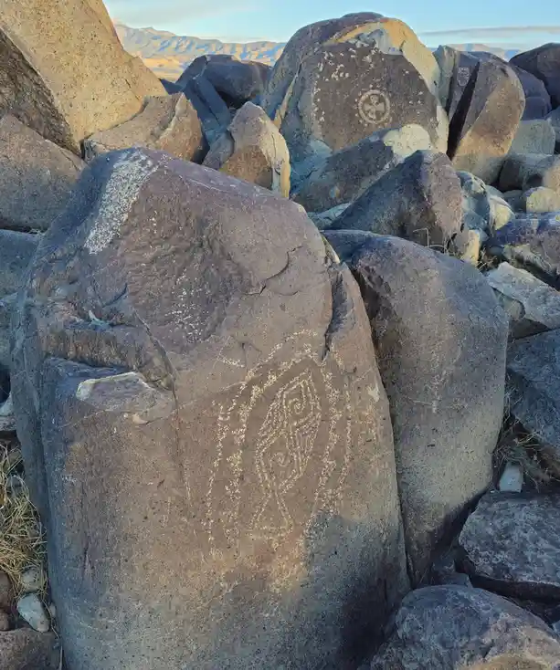 A photo of large, weathered boulders in a desert landscape with ancient petroglyphs carved into their surfaces, including a…