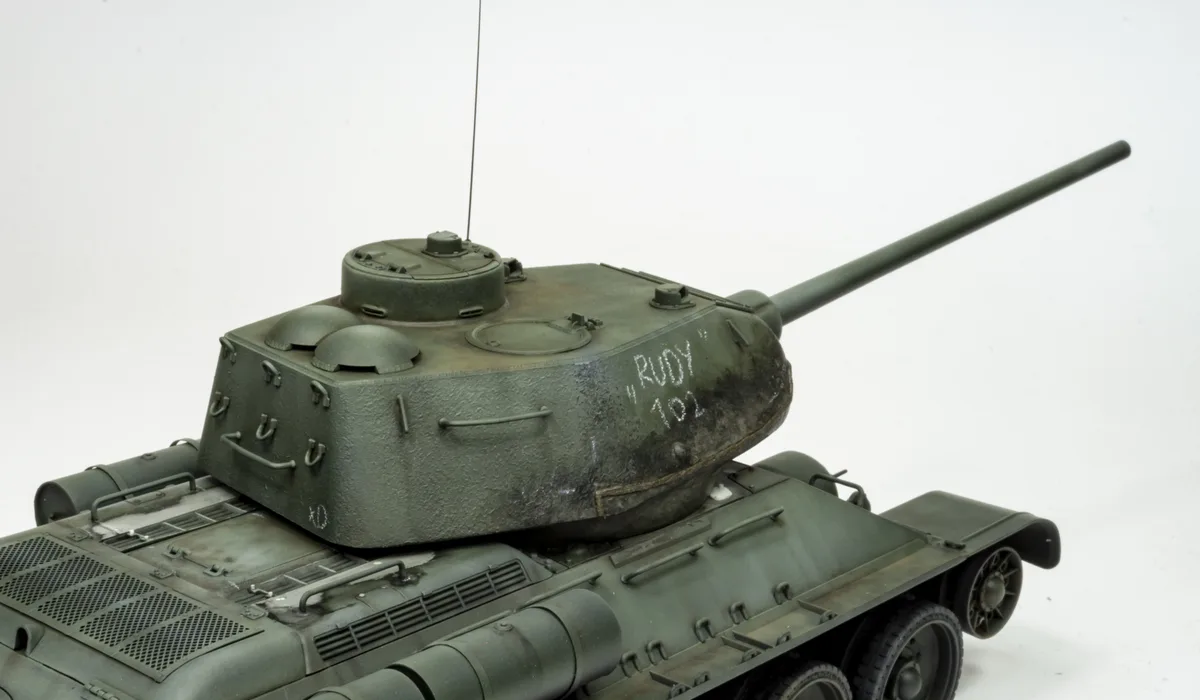 A detailed green military tank model, likely a T-34, is shown against a plain white background. The tank's turret has white…