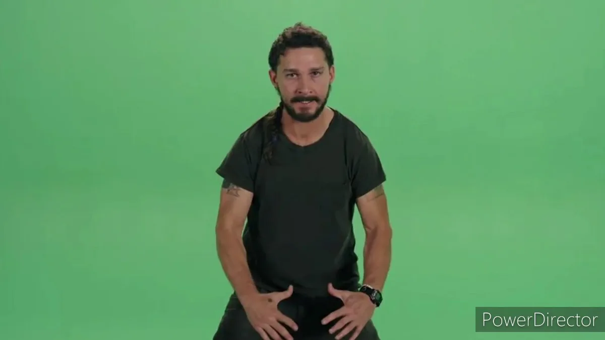 A man with a beard and dark t-shirt stands against a solid green screen background, hands on his hips, looking directly at…