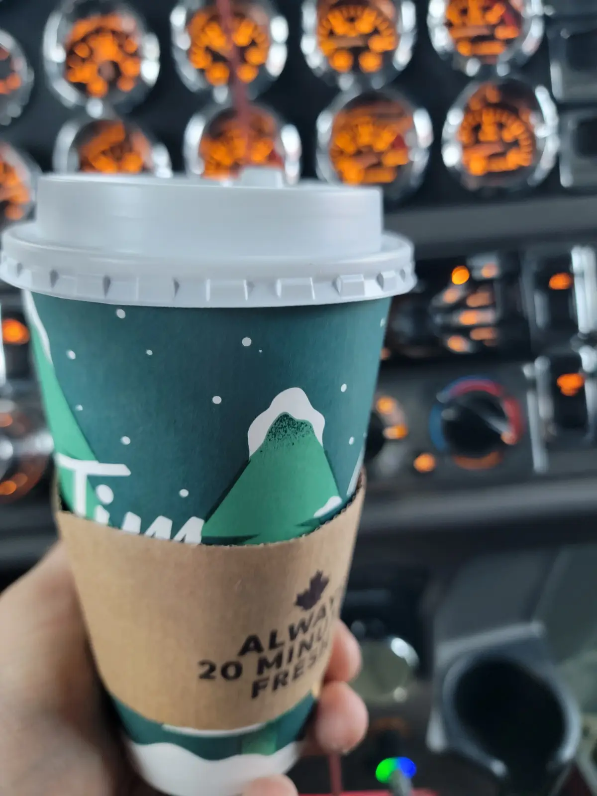 A hand holds a Tim Hortons coffee cup with a green sleeve featuring snow-capped mountains and white dots, inside a brown…