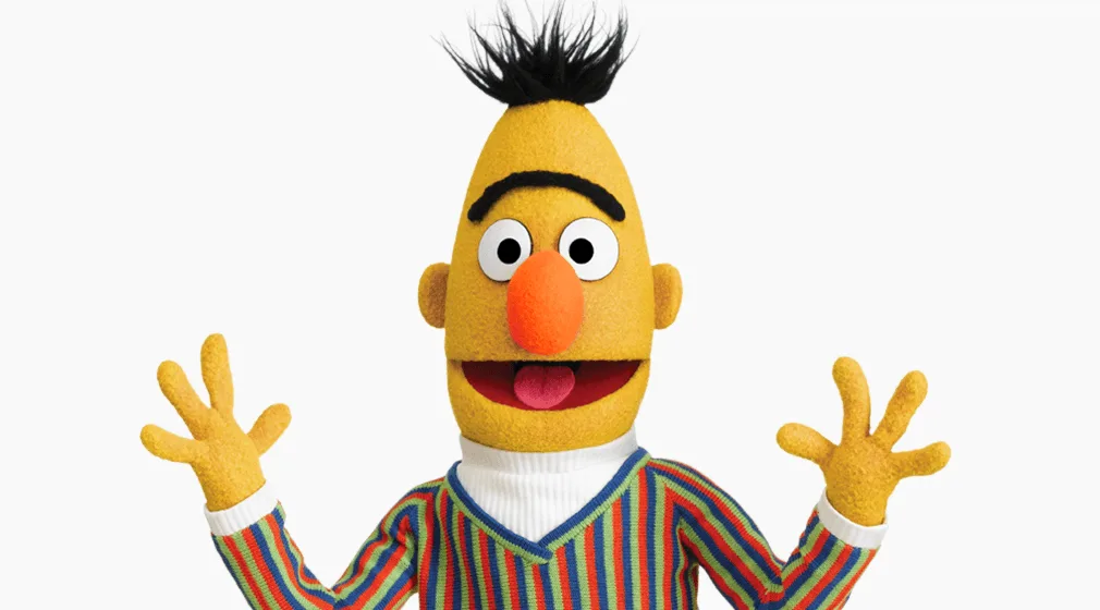 a yellow muppet with a black mohawk, orange nose, and wide eyes wears a striped sweater and raises both hands in a shrugging…