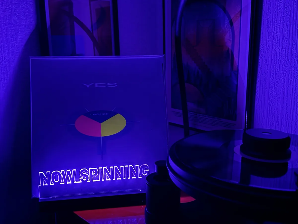 A vinyl record player sits under purple lighting next to a glowing album cover for Yes' '90125' with the text 'NOWLSPINNING'…