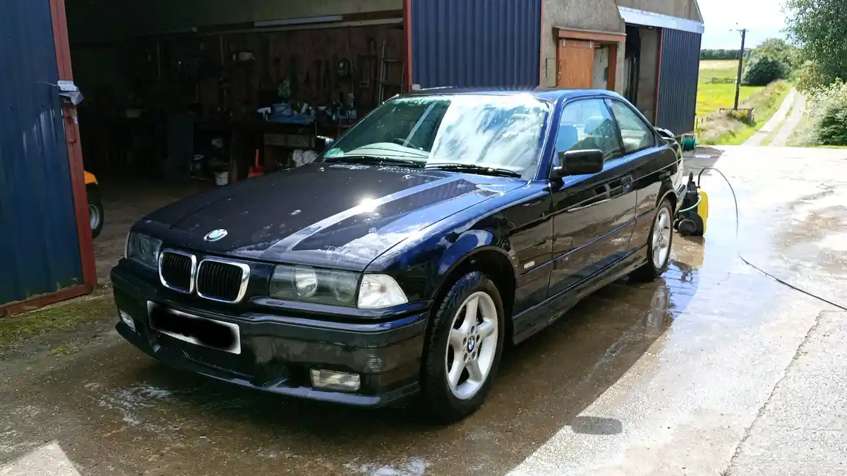 A dark blue BMW E36 coupe is parked outside a garage on a wet driveway, with soap suds visible on its hood and a pressure…