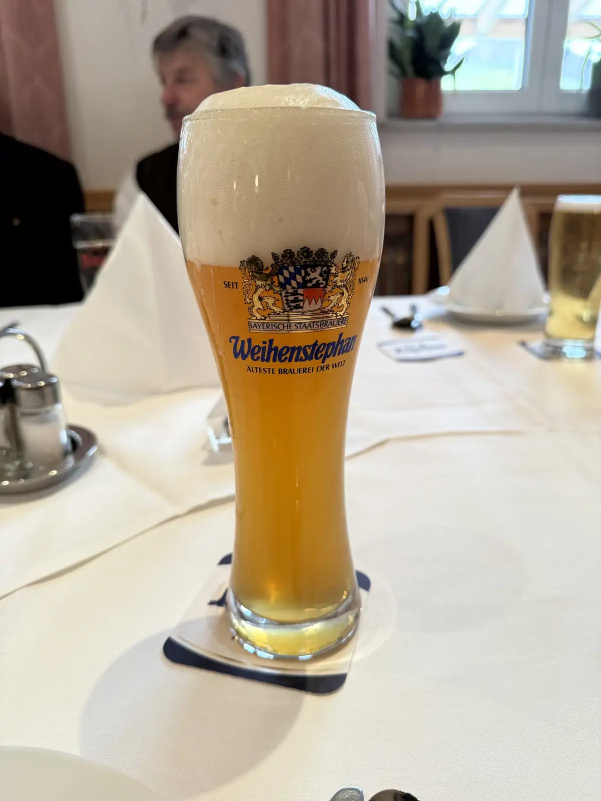 A tall, frothy glass of Weihenstephan beer sits on a coaster at a restaurant table, with the brand's crest and text 'SEIT…