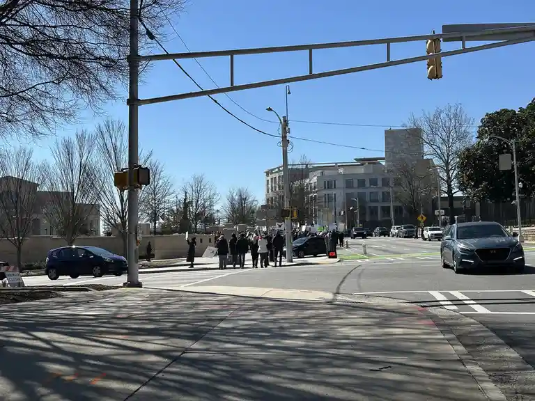 A street-level photo shows a group of protesters holding signs and standing in the middle of a road intersection, partially…