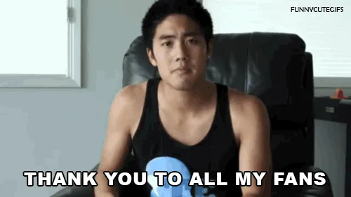 A man in a black tank top sits in a chair and gestures toward two white standing fans and a black tower fan on the floor…
