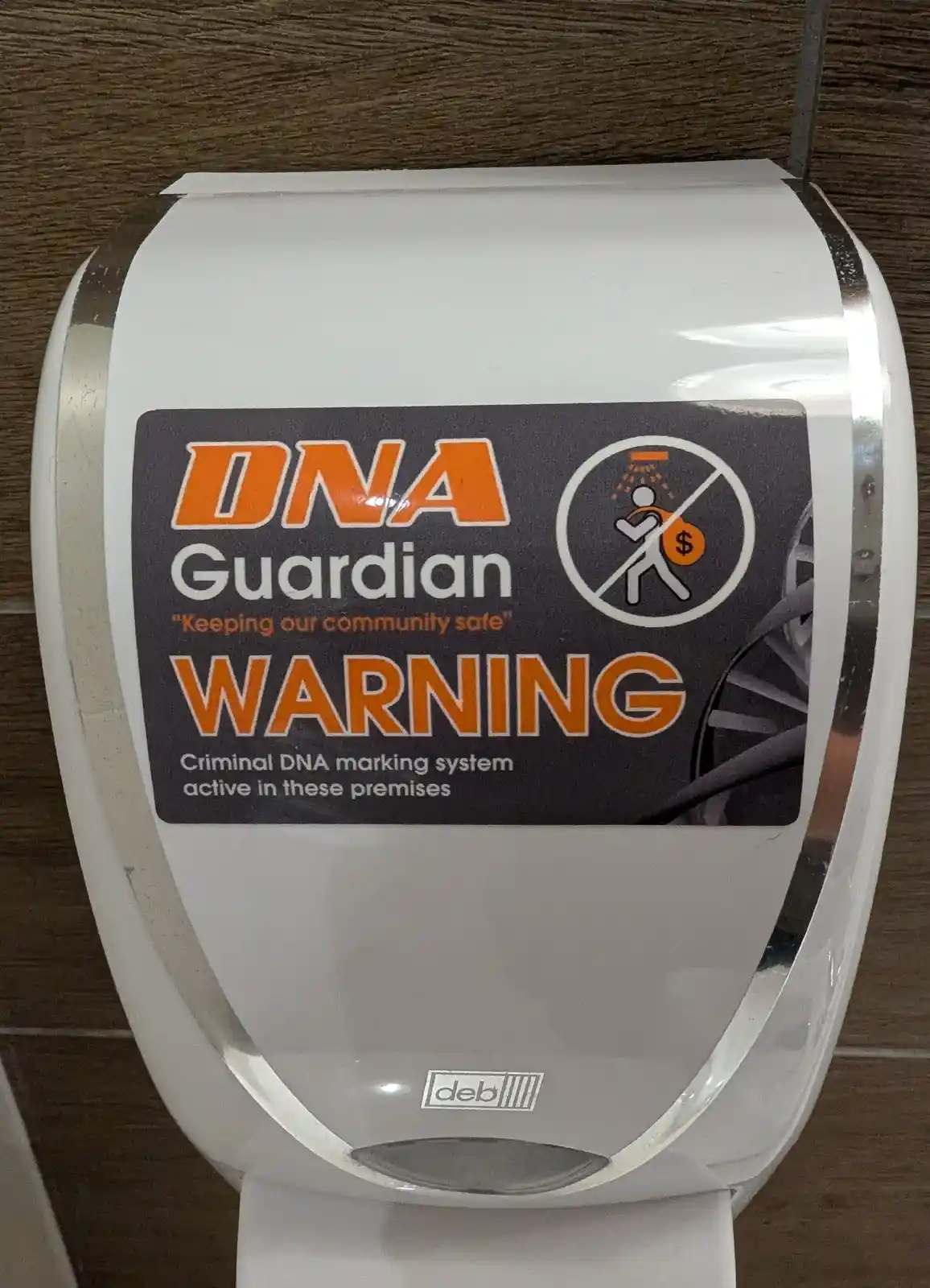 A white, curved soap or sanitizer dispenser mounted on a wood-paneled wall has a large black sticker. The sticker reads 'DNA…