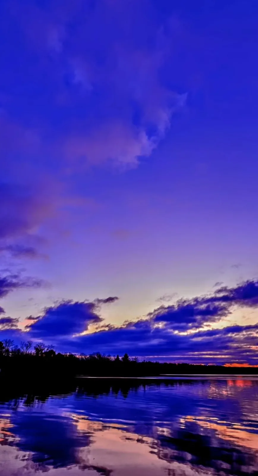 A serene twilight scene over a calm body of water, with a vibrant purple and blue sky reflecting in the water, silhouetted…