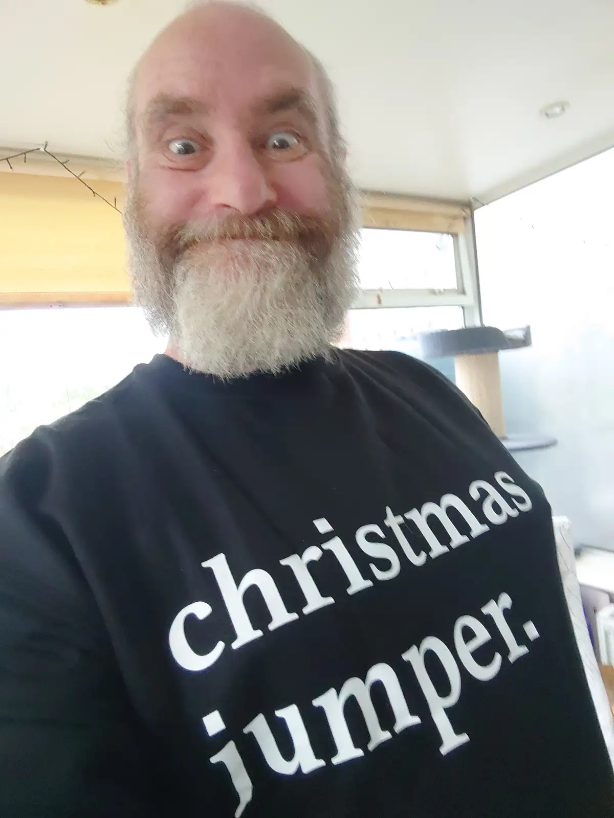 A bald man with a full grey beard and wide, surprised eyes takes a selfie wearing a black t-shirt that reads 'christmas…