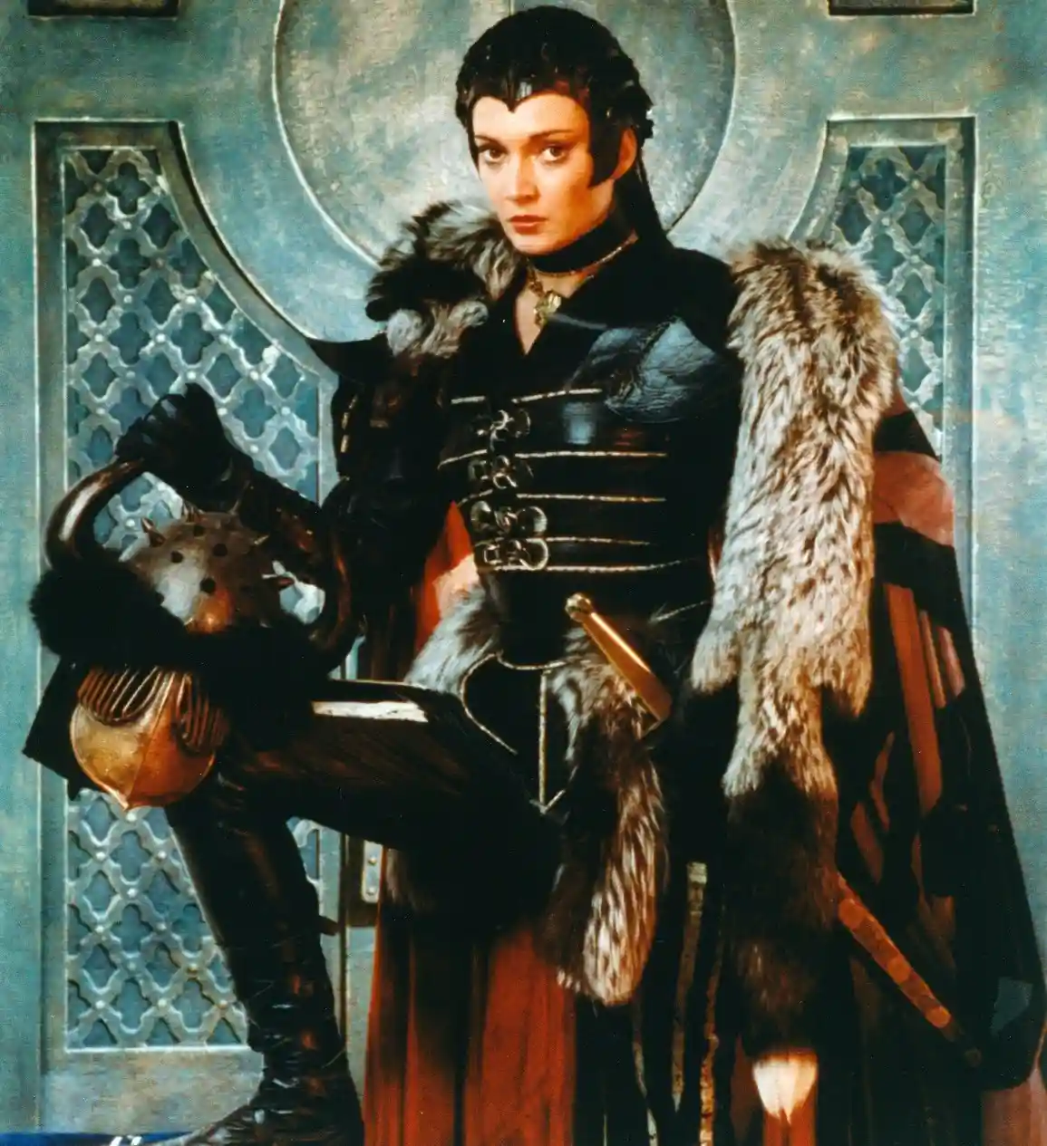 A woman in dark, fur-trimmed medieval-style armor and a red cape stands in front of an ornate blue door, holding a spiked…